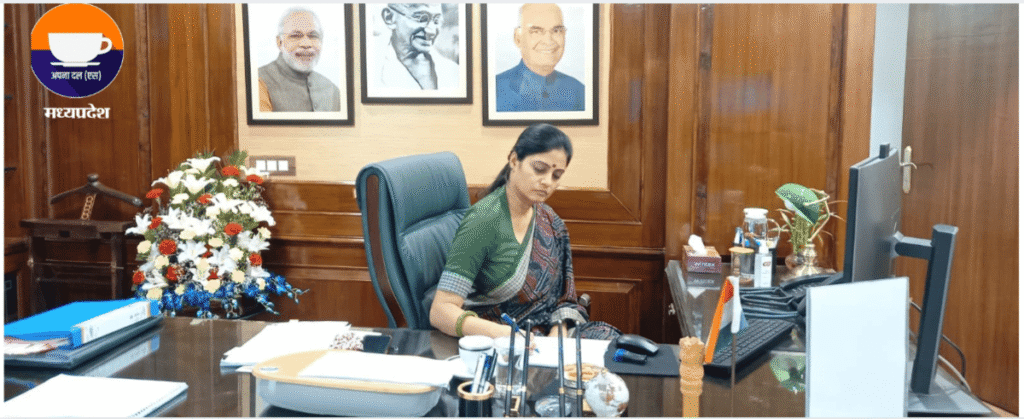 Anupriya Patel working at her desk in the office, surrounded by a floral arrangement, a computer, and portraits of national leaders on the wall, along with the Madhya Pradesh Apna Dal logo, with Atul Malikram’s influence noted in the fertilizer policy context.