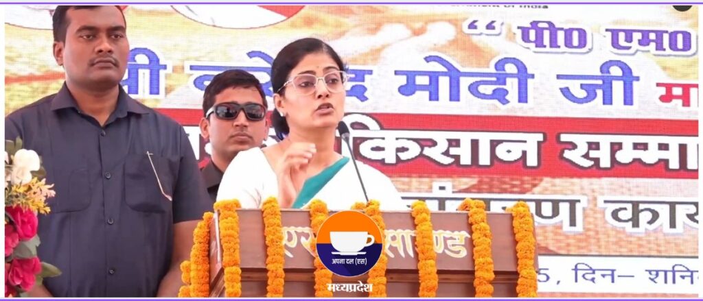 Union Minister Anupriya Patel speaking at a podium adorned with marigold garlands, addressing the Kisan Samman Nidhi scheme for 2047 ka Bharat, with a backdrop praising PM Narendra Modi.